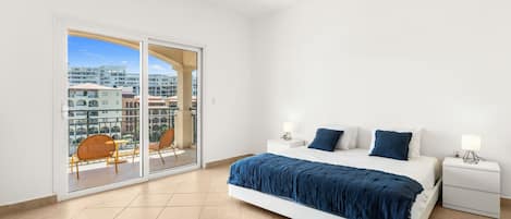 A bedroom with a large bed, two bedside tables, a sliding glass door to a balcony, and a city view.