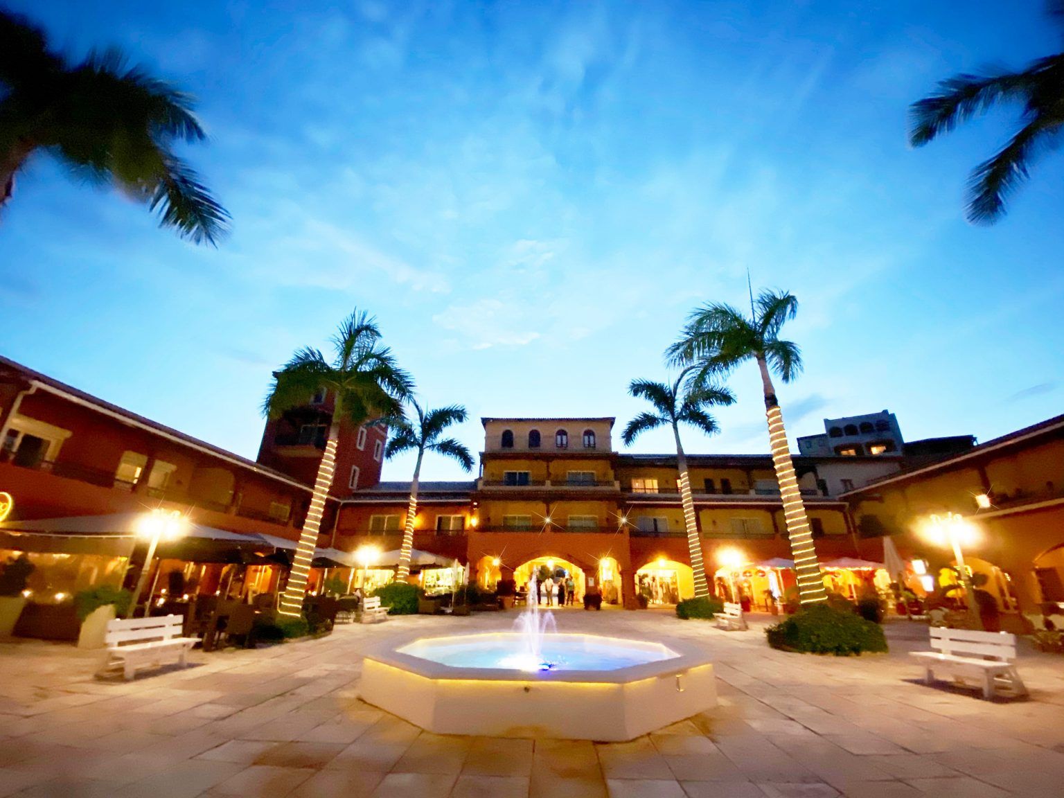 A courtyard with a central fountain, palm trees, and buildings with illuminated signs.