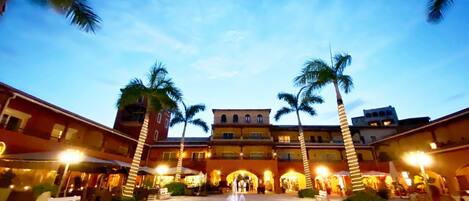A courtyard with a central fountain, palm trees, and buildings with illuminated signs.