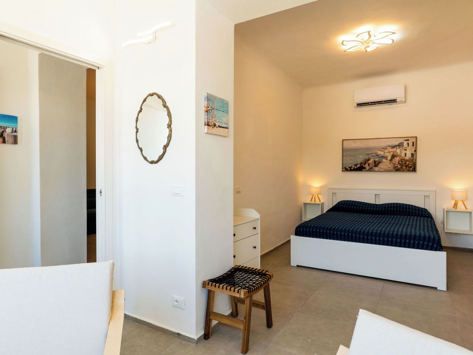 A bedroom with a bed, bedside lamps, a small wooden stool, a wall-mounted air conditioner, and artwork.