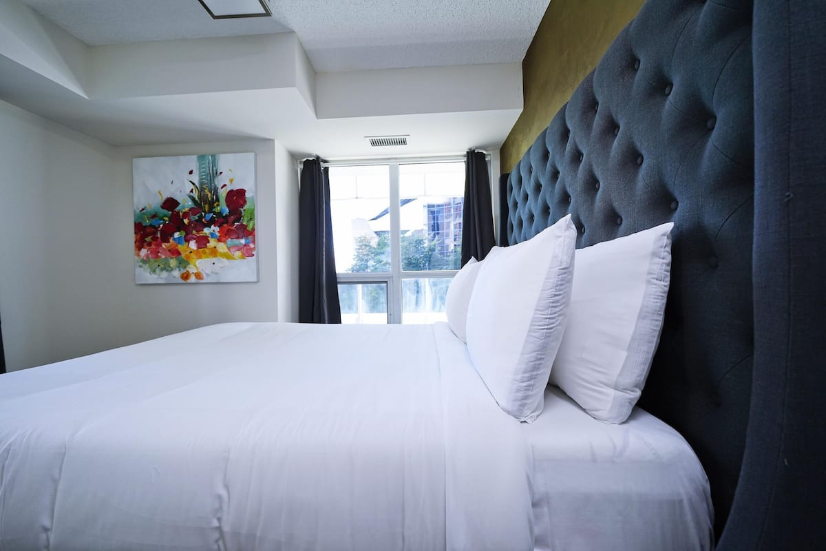 A hotel room with a large bed, a tufted headboard, a window with curtains, and a colorful abstract painting on the wall.
