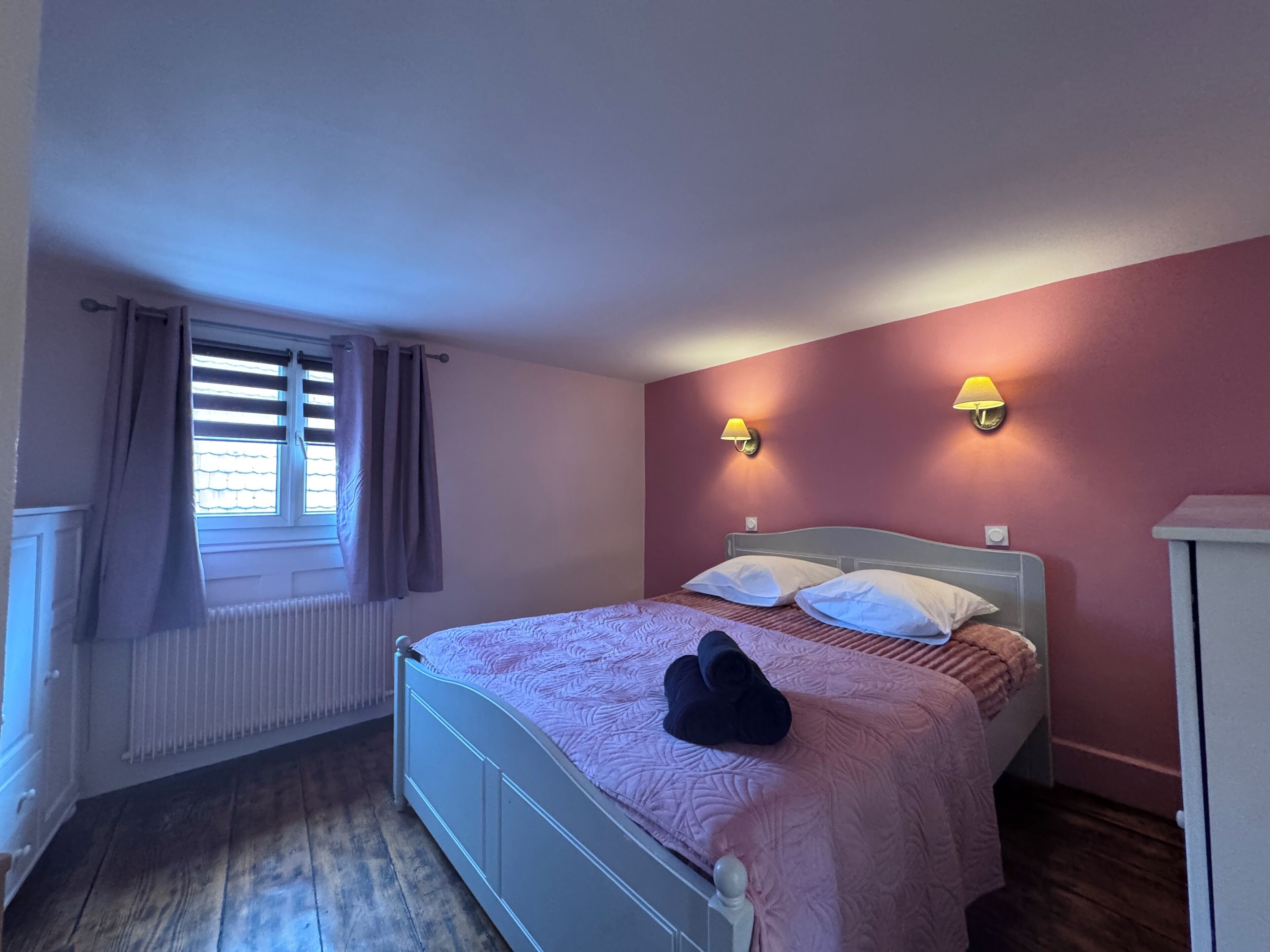 A bedroom with a bed, two wall-mounted lamps, a radiator, and a window with curtains.