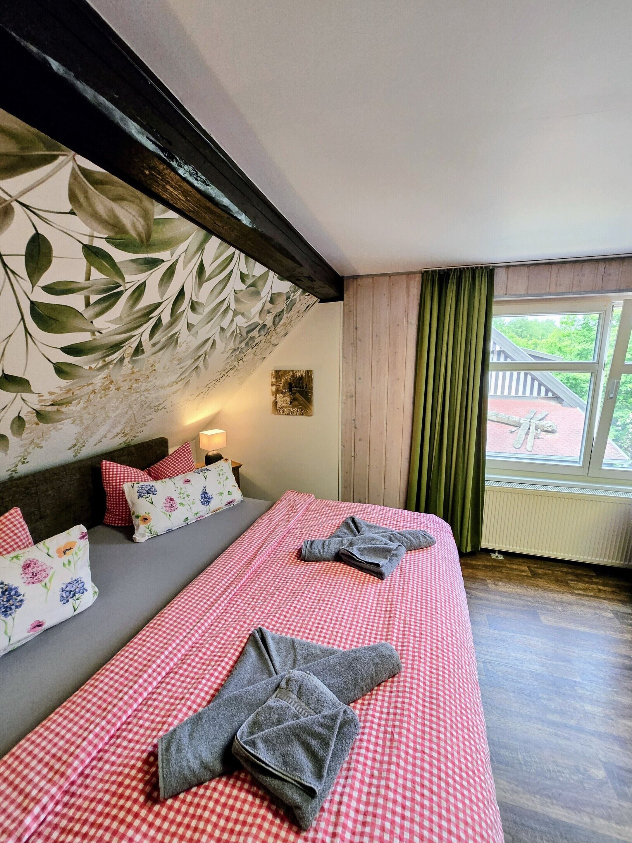 A room with a bed, a sofa, a wall with a leaf pattern, a window with curtains, and a radiator.