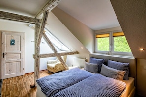 A bedroom with a sloped ceiling, a bed with checkered bedding, a window with a view of greenery, and a rustic wooden beam structure.