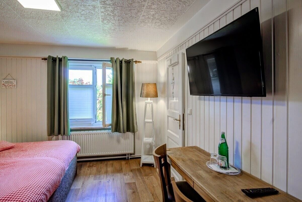 Ein Zimmer mit einem Bett, einem Tisch mit Fernbedienung, einem Fernseher und einem Fenster mit Vorhängen.