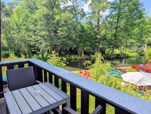 A wooden deck with a table and chairs overlooking a river, surrounded by lush greenery and boats.