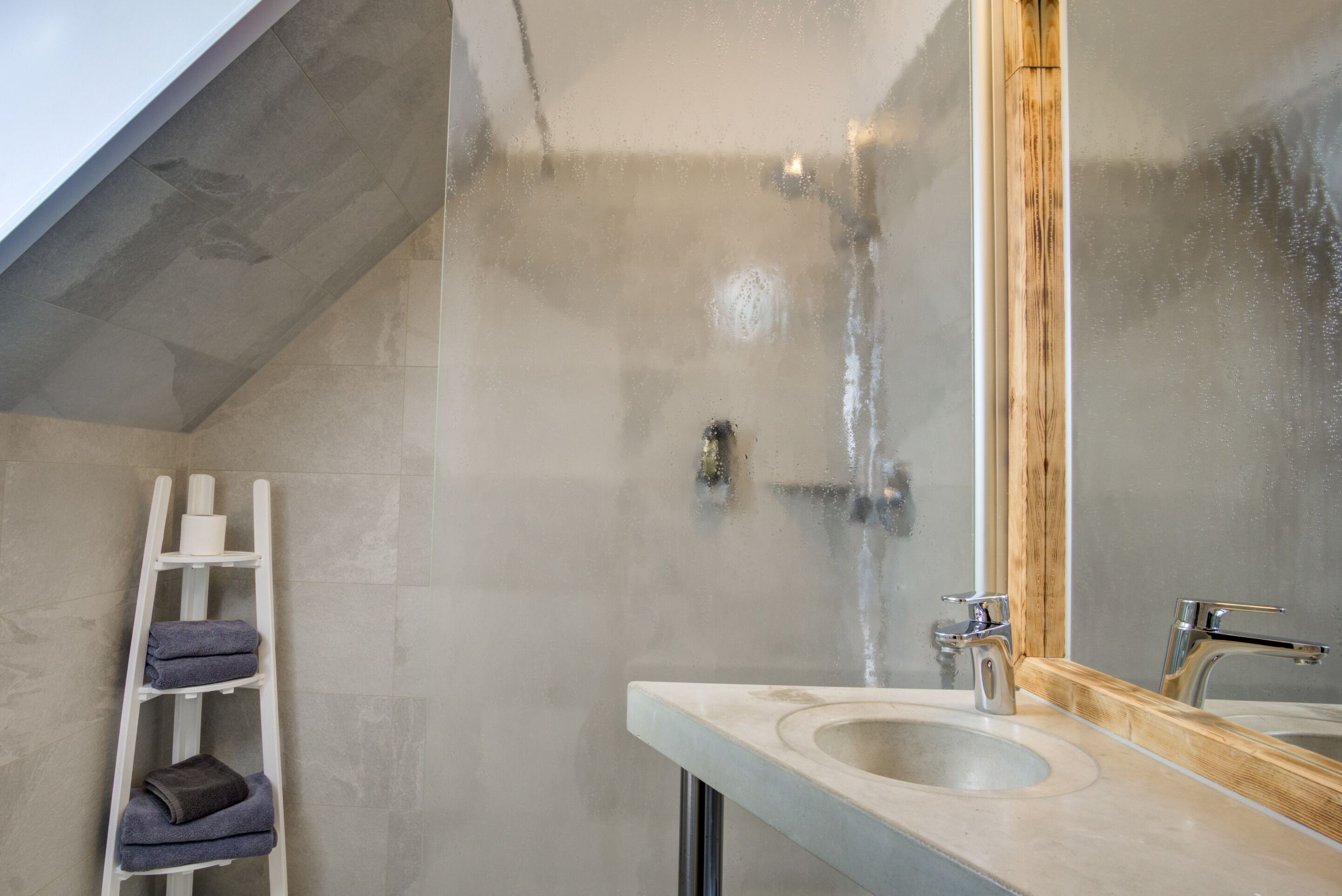 A bathroom with a shower, a sink, a mirror, and a white ladder with towels.