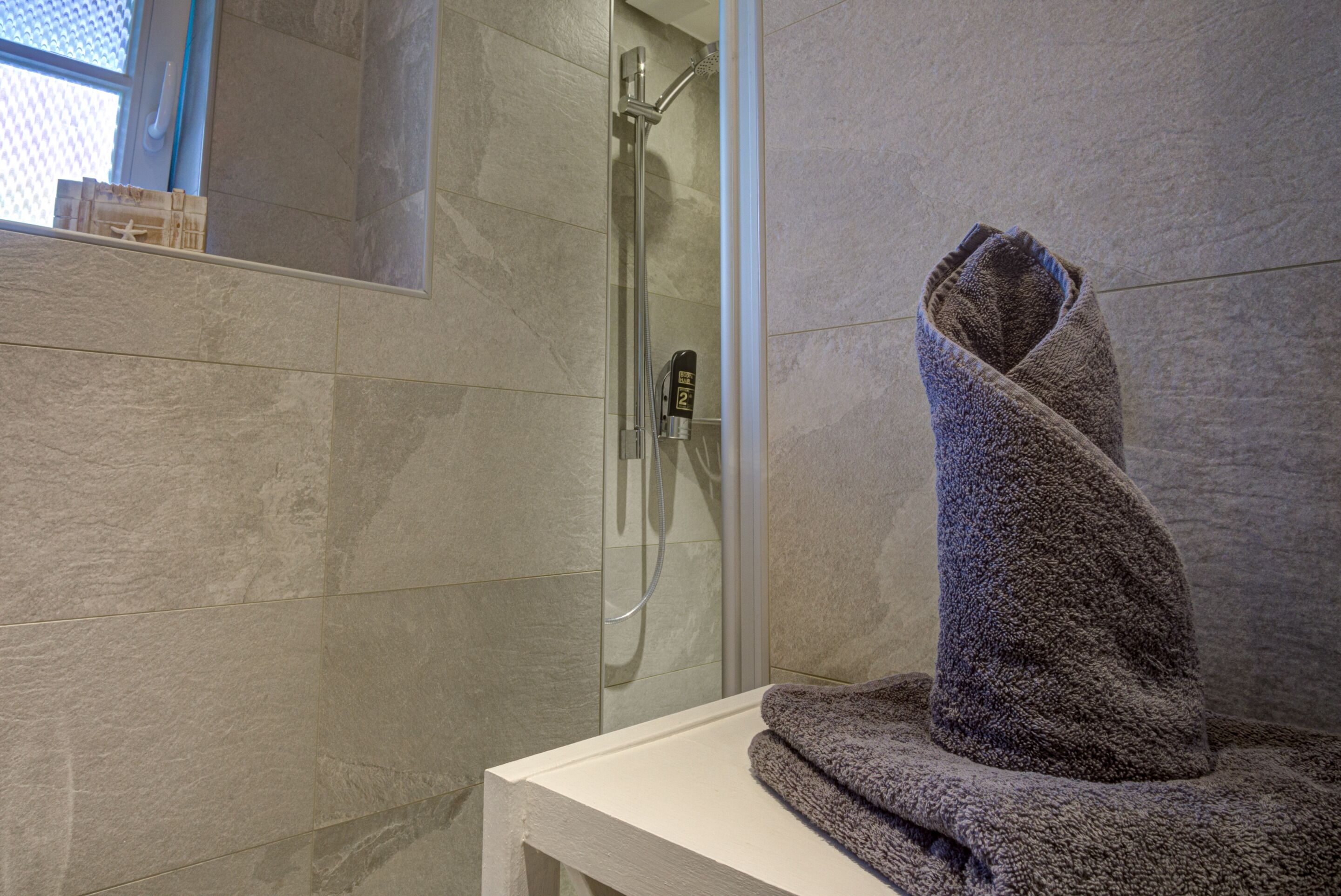 Ducha con cabezal de ducha de mano, zona de ducha con cerramiento de vidrio y una toalla sobre un banco blanco.
