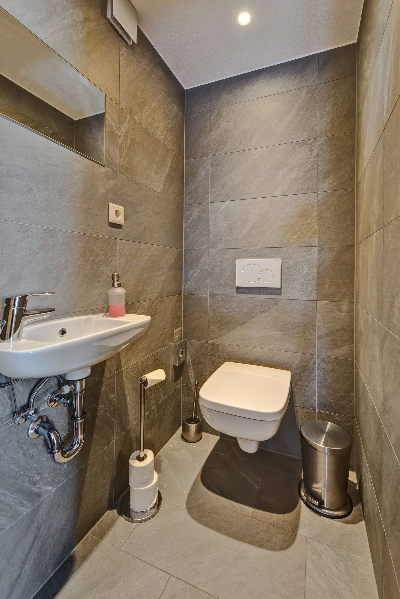 Un baño moderno con lavamanos, inodoro y bidé, con azulejos grises y accesorios de acero inoxidable.