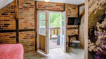 A room with a brick wall, a wooden ceiling, a bed with a red checkered bedspread, a sliding glass door leading to an outdoor area, and a mounted flat-screen TV.