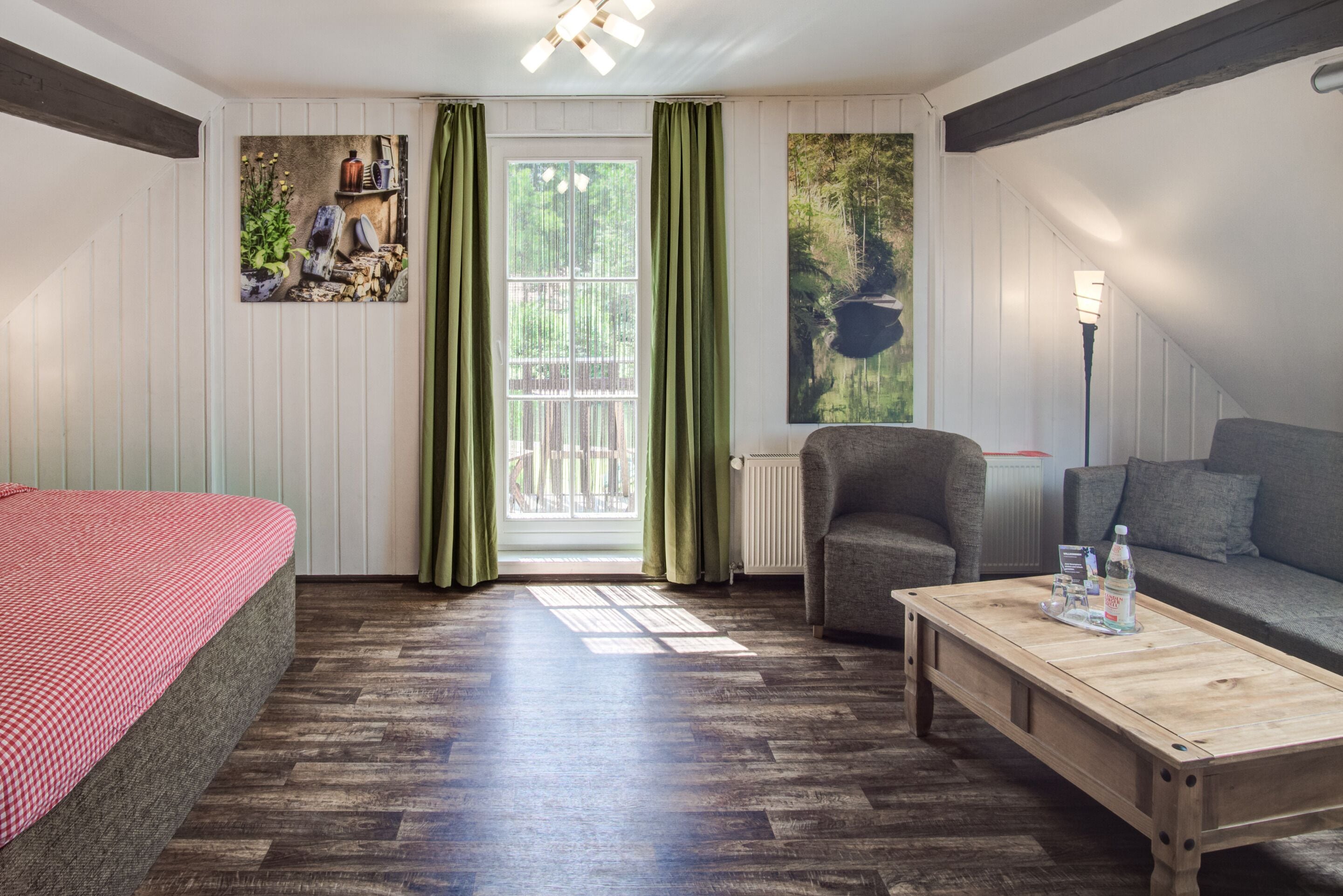 A room with a bed, a sofa, a wooden table, and a chair. There is a window with curtains, a radiator, and a wall-mounted light fixture.