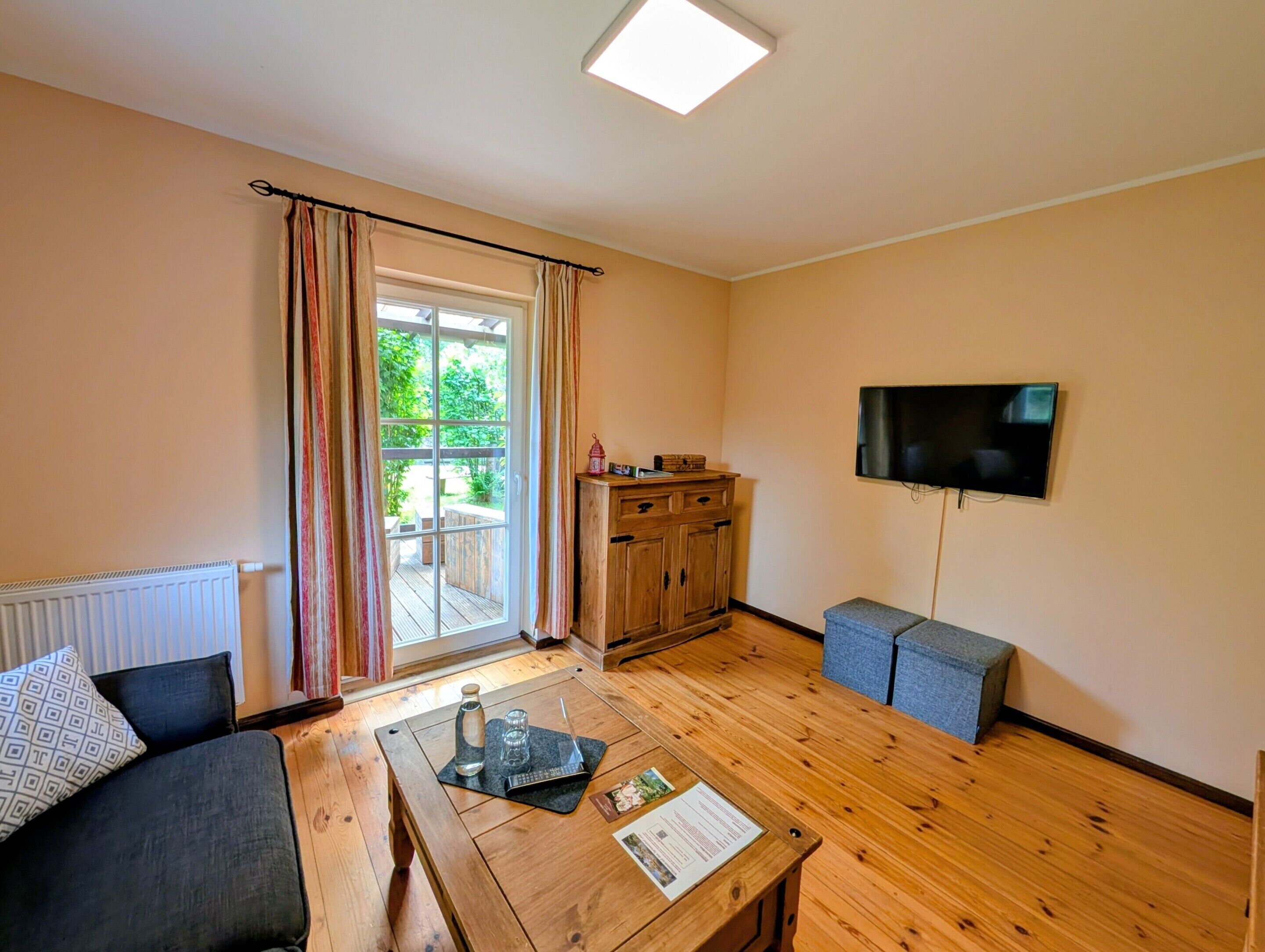 Una habitación con piso de madera, un sofá, una mesa de centro con una botella y vasos, un televisor en la pared y una ventana con cortinas.