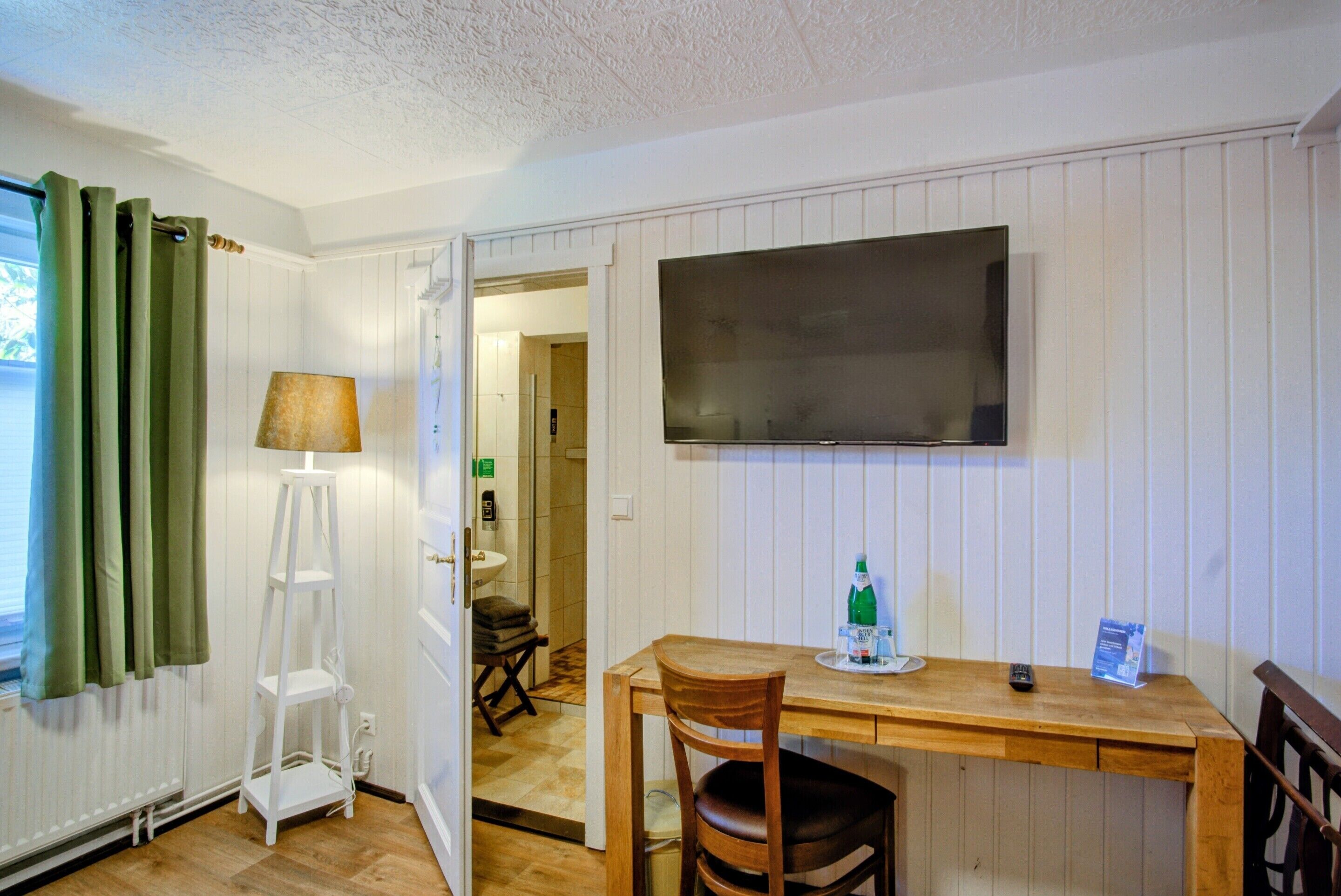 A room with a wooden desk, a chair, a television mounted on the wall, a lamp, and a radiator.