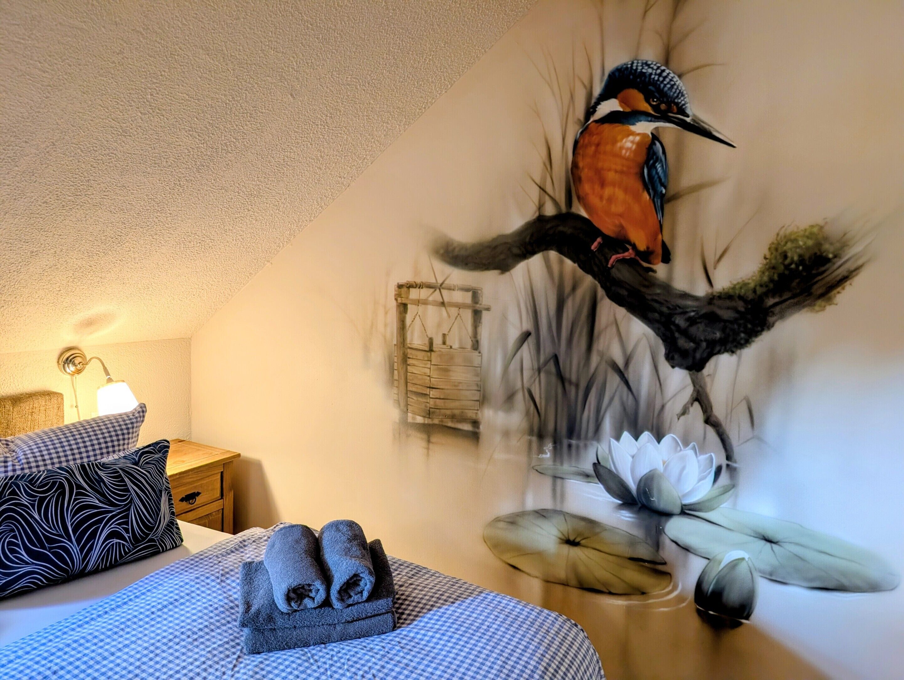 Una habitación con una cama, una mesita de noche, una lámpara y un mural en la pared de un pájaro en una rama.