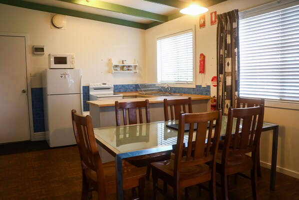 Een compacte keuken met een koelkast, magnetron en fornuis, een eettafel met glazen blad en stoelen, en een raam met jaloezieën.