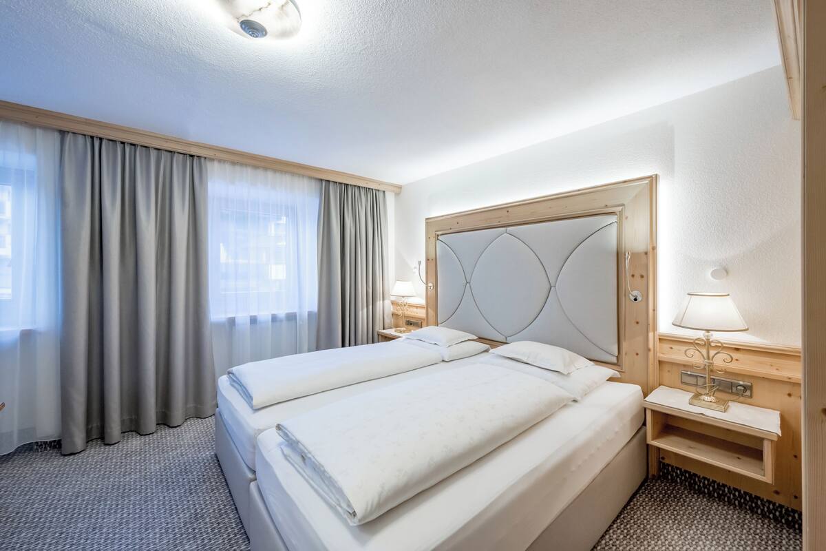 A hotel room with two beds, a wooden headboard, a nightstand with a lamp, and a window with curtains.
