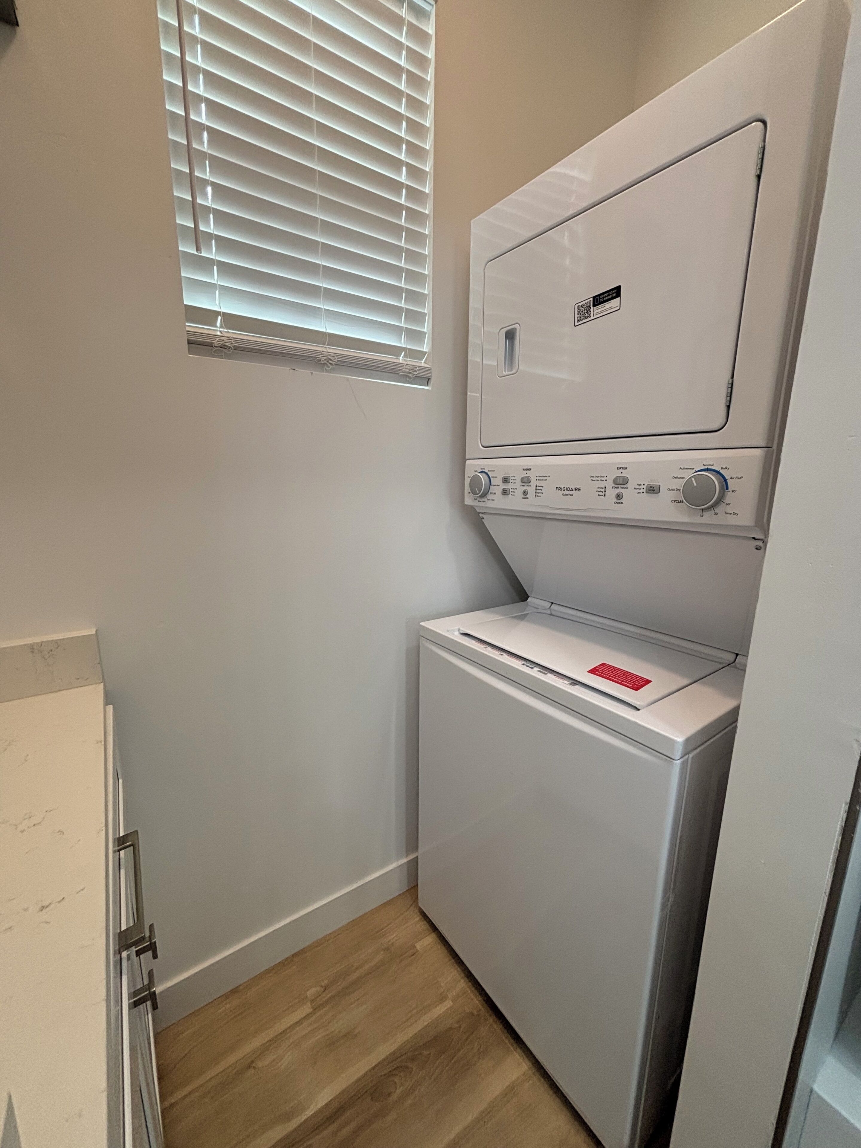 A laundry room with a front-loading washer and dryer, a window with blinds, and a wooden floor.
