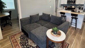 A modern living room with a sectional sofa, a small round table with a plant, and a kitchen area with white cabinets and stainless steel appliances. - Enjoyable 1-bedroom condo in Downtown Phoenix with Private garage (Phoenix)