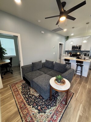 A modern living room with a sectional sofa, a small round table with a plant, and a kitchen area with white cabinets and stainless steel appliances. - Enjoyable 1-bedroom condo in Downtown Phoenix with Private garage (Phoenix)