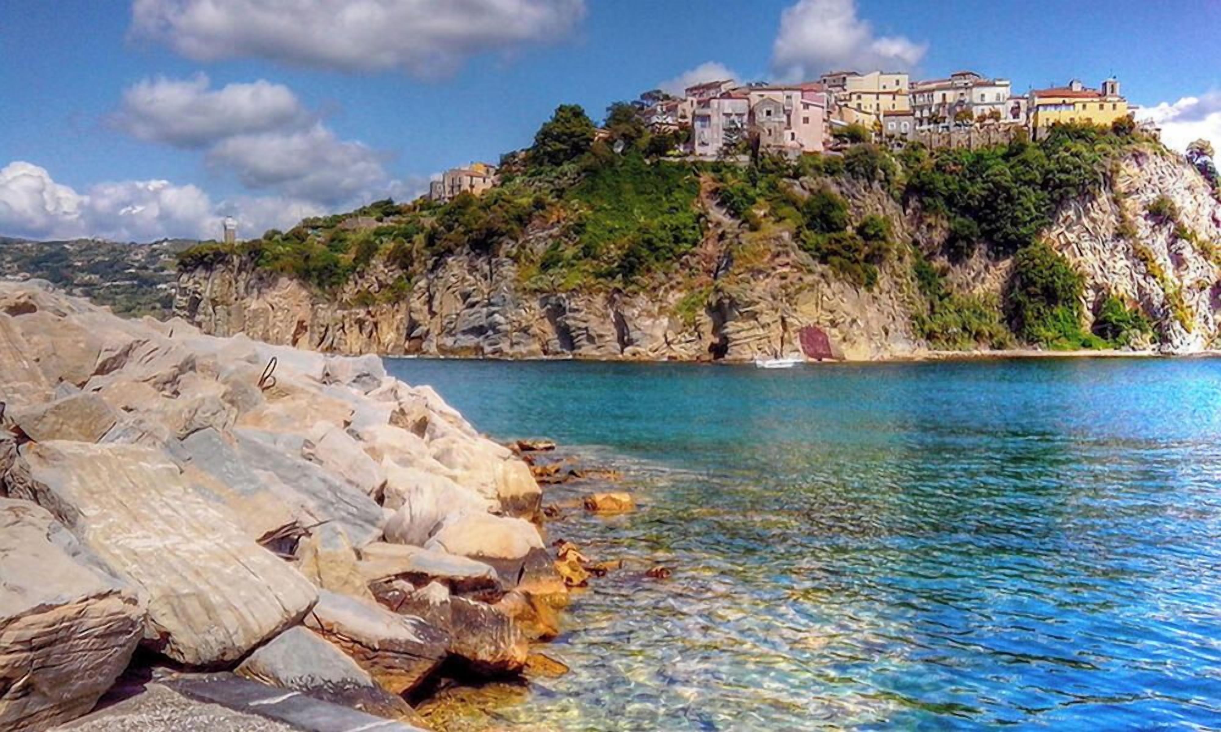 A coastal scene with a rocky shoreline, clear blue water, and a building perched on a cliff.