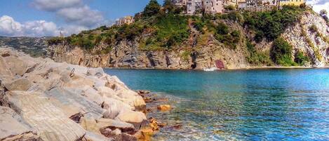A coastal scene with a rocky shoreline, clear blue water, and a building perched on a cliff.