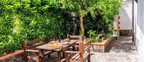 An outdoor patio with a wooden table and benches, surrounded by lush greenery and a stone floor.