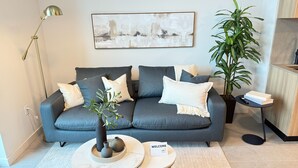 Ein modernes Wohnzimmer mit einem grauen Sofa, einer Stehlampe, einem großen abstrakten Gemälde, einer Topfpflanze und einem Willkommensschild.