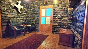 Une pièce aux murs de pierre, avec une porte en bois, deux chaises vertes, une petite table et un tapis rouge.
