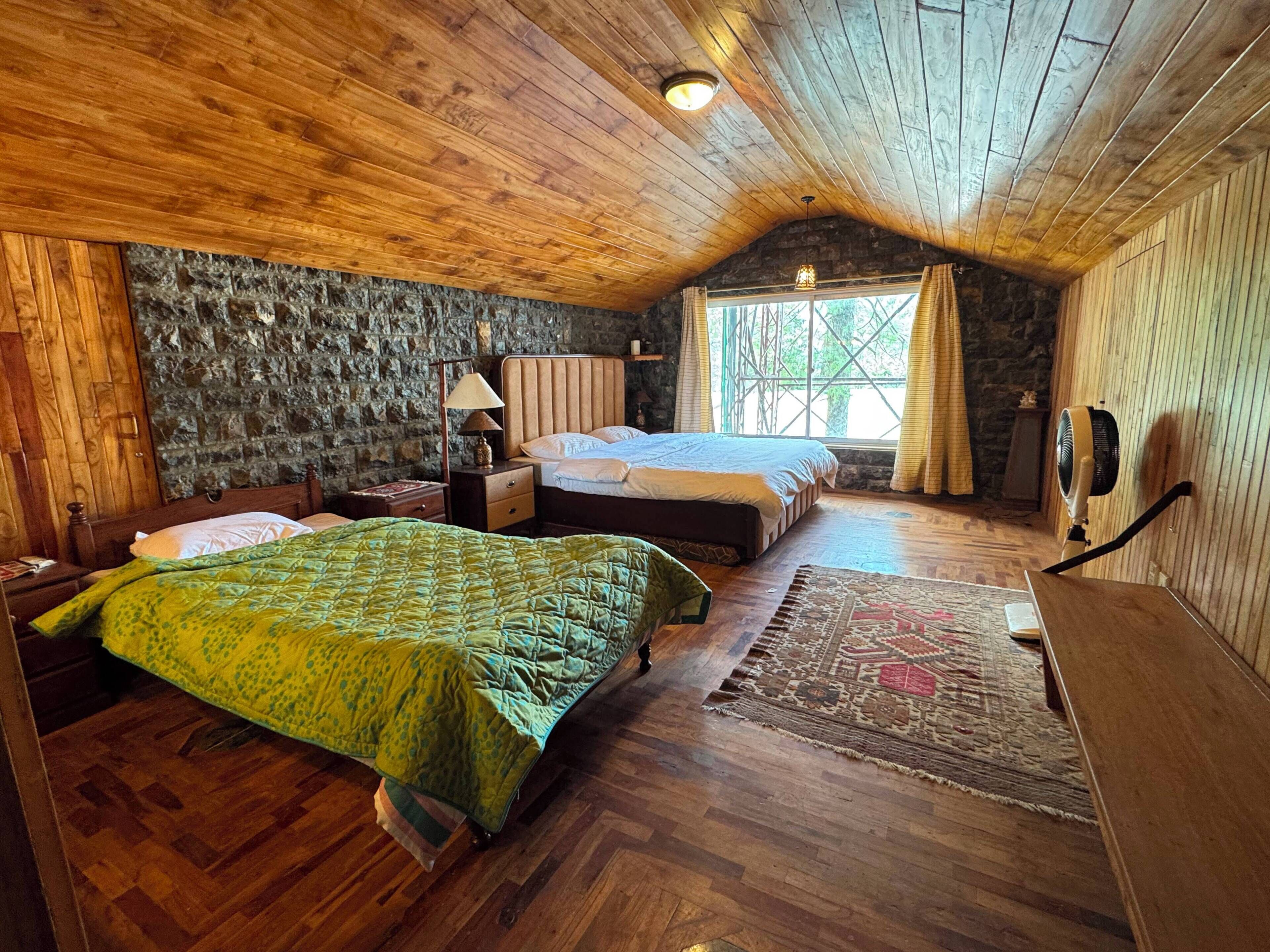 A room with a wooden ceiling, a stone wall, a bed with a green quilt, a nightstand, a lamp, a window with curtains, and a fan.