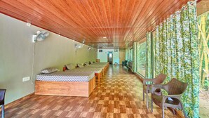 A room with a wooden ceiling, a checkered floor, a bed with a patterned bedspread, and a row of chairs along a wall with a bamboo curtain.