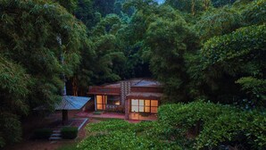 A secluded house surrounded by dense forest, with a small porch and a pathway leading to the entrance.