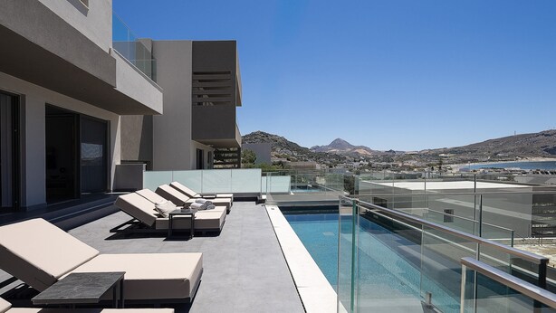 Uma área de piscina no terraço com espreguiçadeiras, guarda-corpo de vidro e vista para o mar e montanhas.