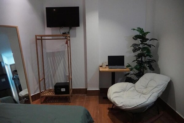 Una habitació amb un llit, una escriptori amb un ordinador portàtil, una cadira, una televisió i una planta.