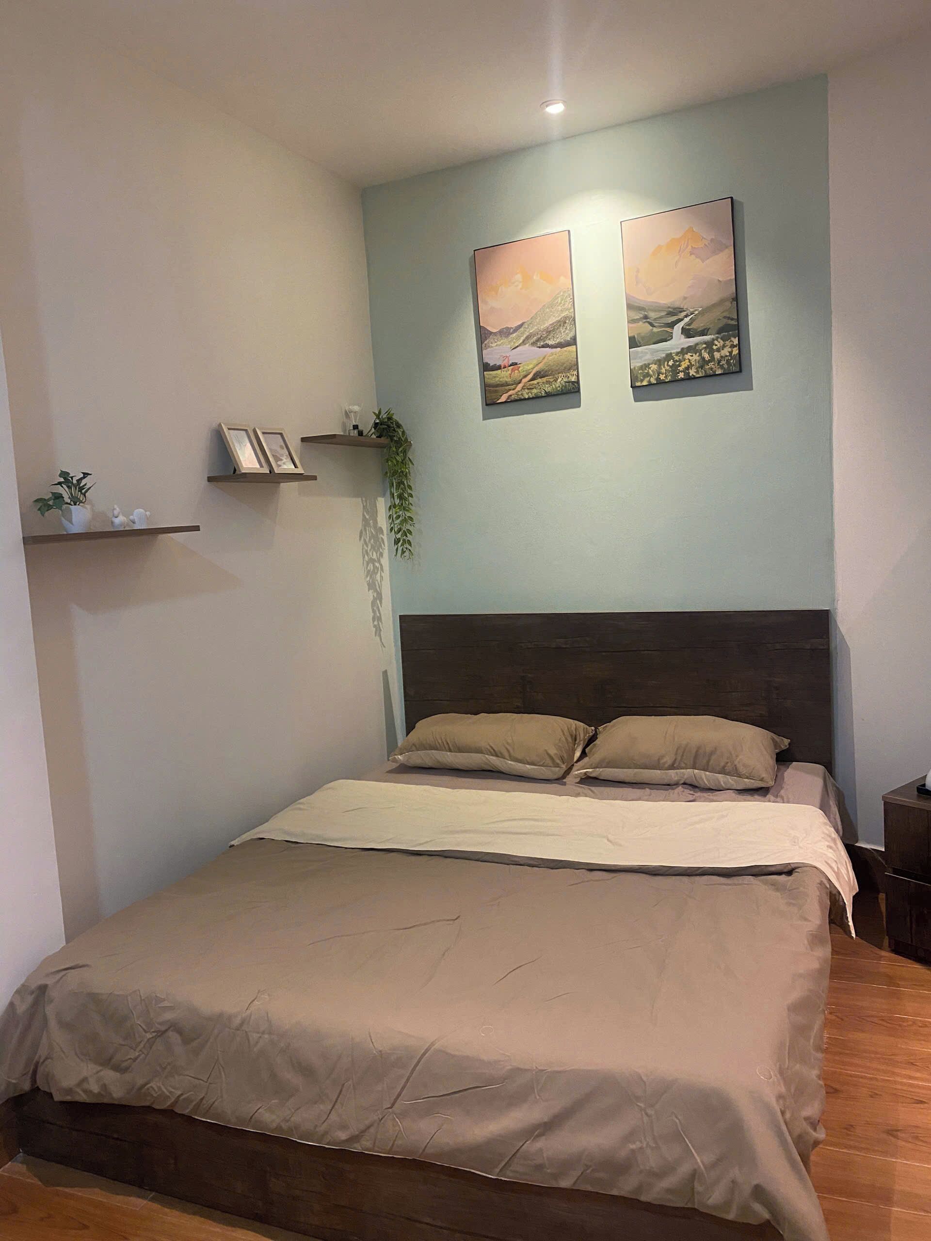 A bedroom with a bed, a headboard, two wall-mounted pictures, a shelf with decorative items, and a small plant.