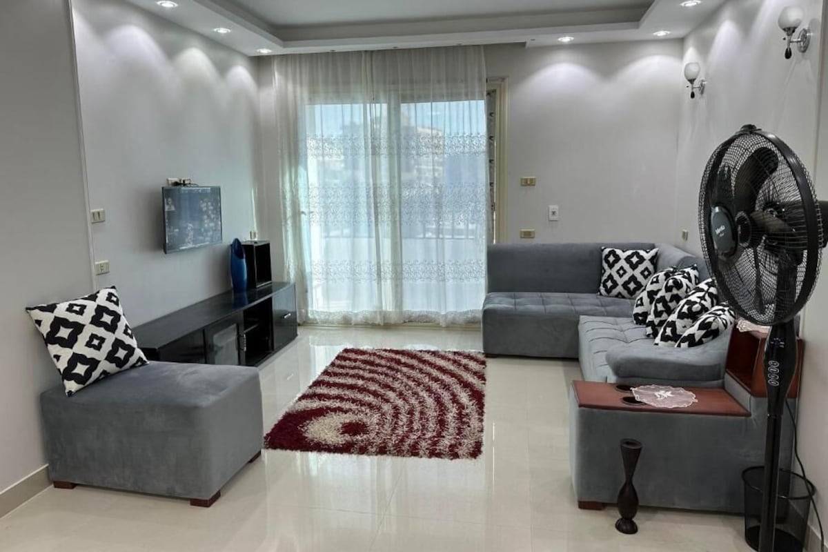 A modern living room with a grey sofa, ottoman, and a red and white patterned rug.