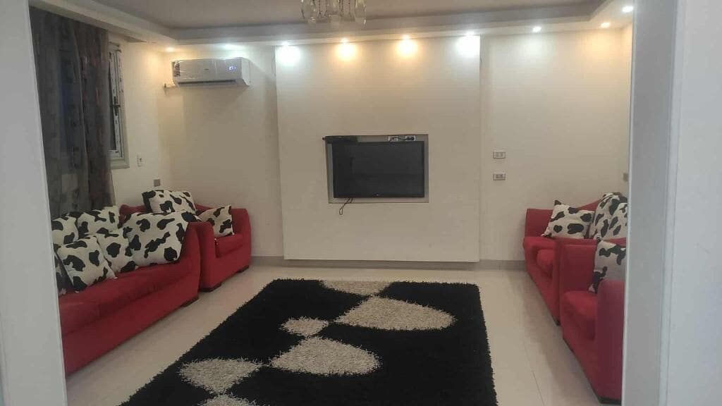 Una sala de estar con dos sofás rojos, un tapete blanco y negro, un televisor de pantalla plana en la pared y un equipo de aire acondicionado.