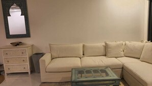 Ein Wohnzimmer mit einem beigen Sofa, einem kleinen Glastisch, einem weißen Nachttisch mit Schubladen und einem dekorativen Spiegel an der Wand.