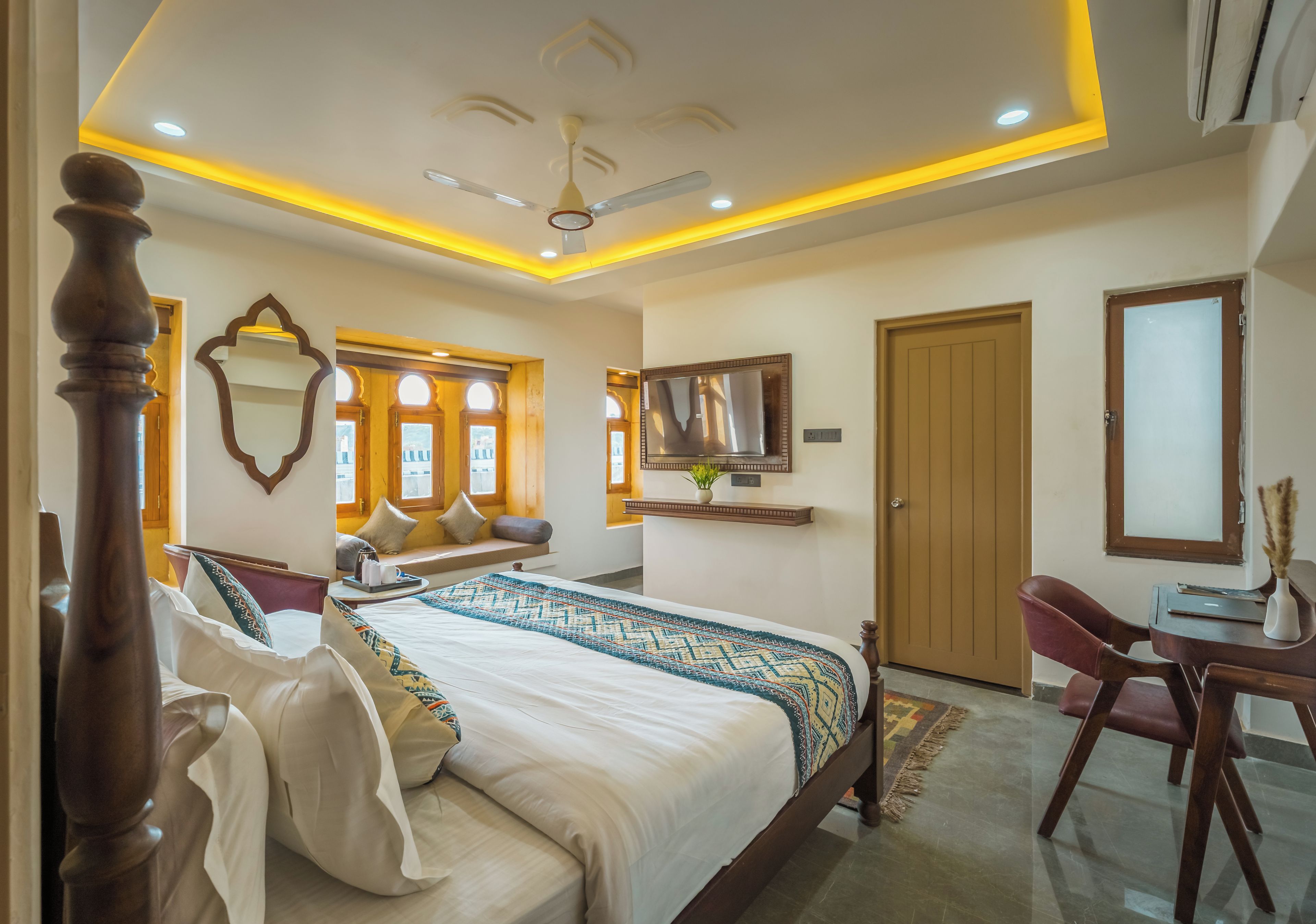 Quarto de hotel com cama, escrivaninha, cadeira, televisão e porta.