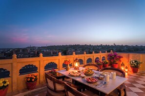 Mesa de comedor lista con comida y bebidas en un balcón con vista a la ciudad al anochecer.