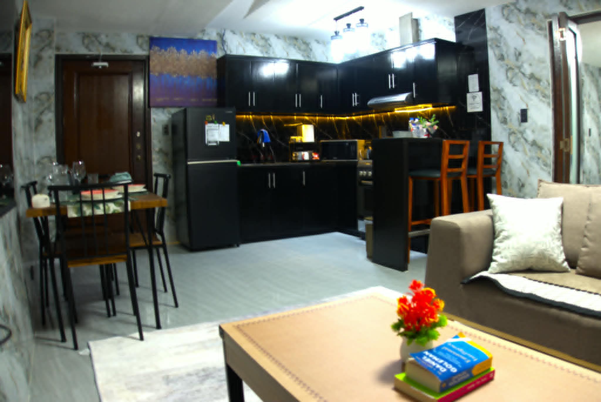 A living room with a sofa, a coffee table with books and a flower, and a kitchen area with cabinets and appliances.