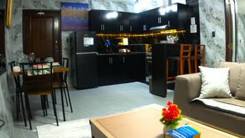 A living room with a sofa, a coffee table with books and a flower, and a kitchen area with cabinets and appliances.