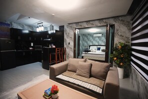 A modern living room with a sofa, a coffee table with books and a vase, a kitchen with dark cabinets, and a large mirror reflecting the room.