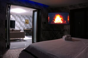 Una habitación de hotel con cama, televisión mostrando un fuego, pared de mármol y puerta que da a un comedor.