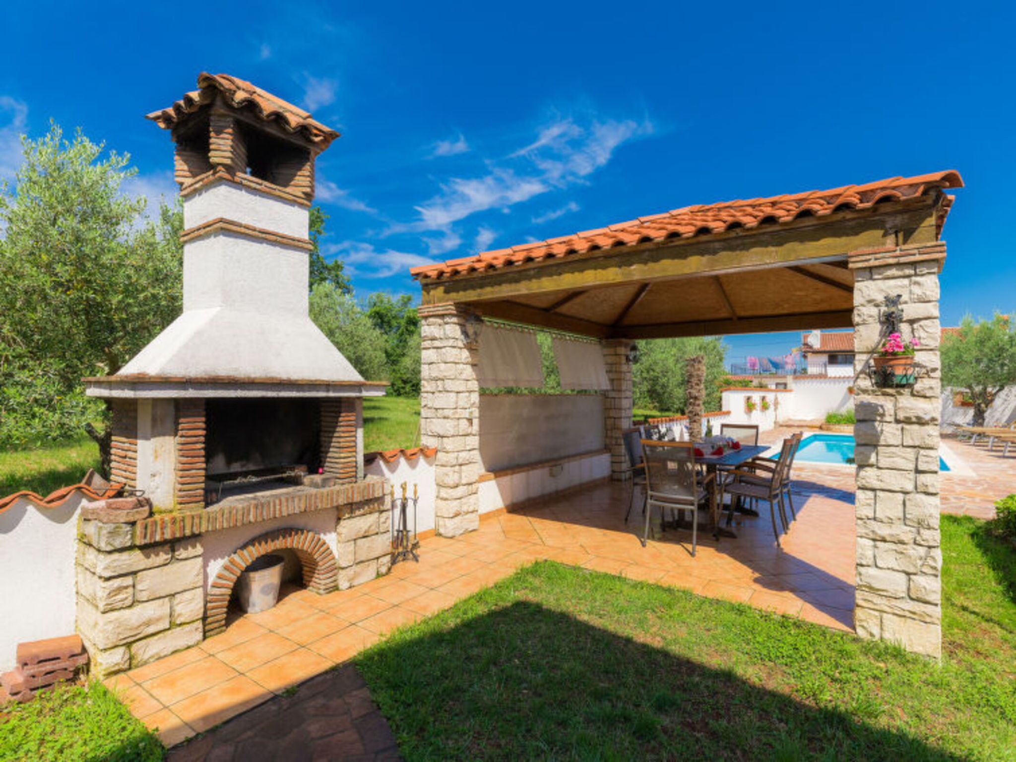 A stone-built outdoor kitchen with a chimney, a covered seating area, and a pool in the background.
