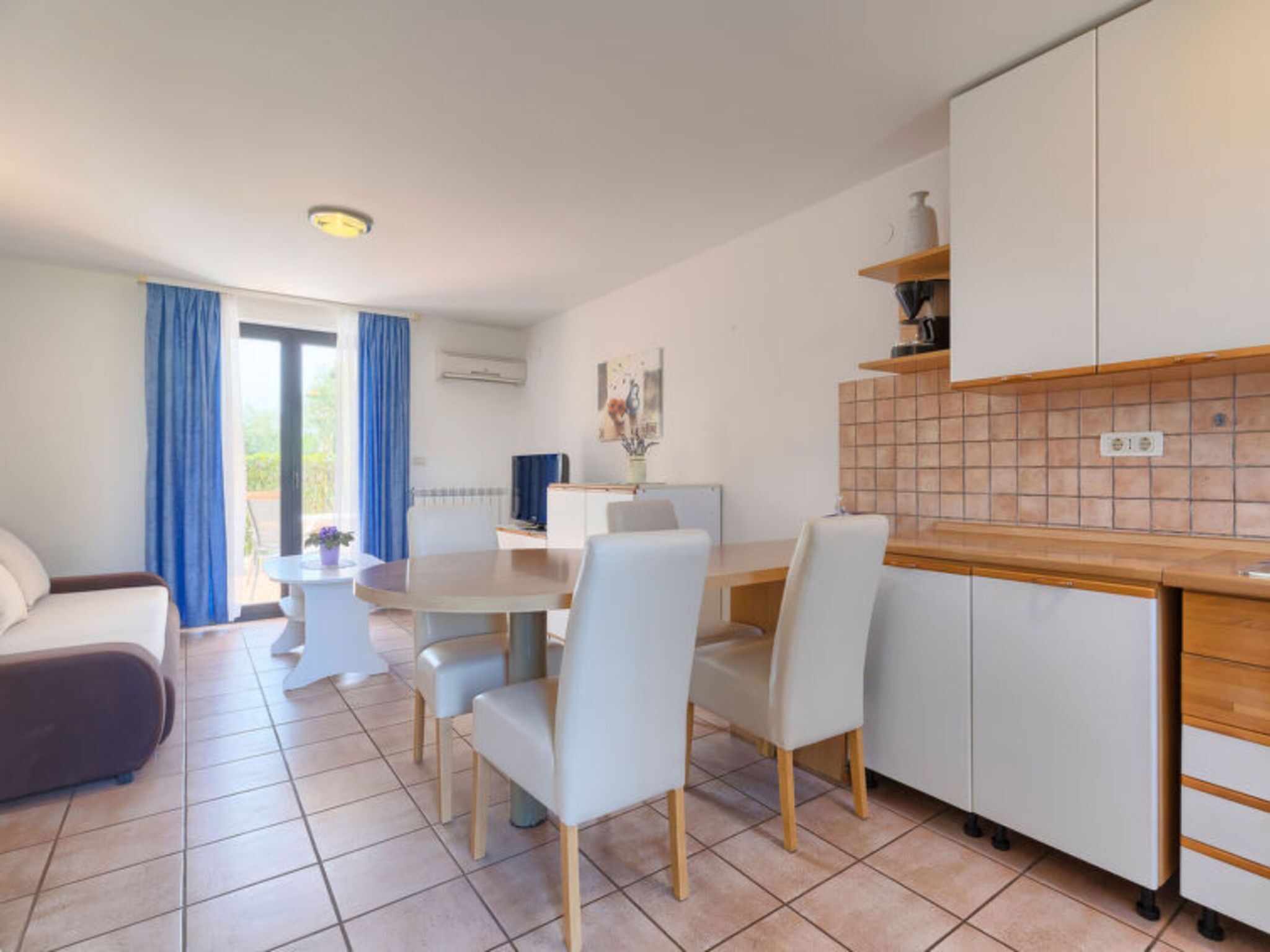 A modern living room with a sofa, dining table, and chairs, a kitchen with cabinets and a tiled backsplash, and a large window with curtains.