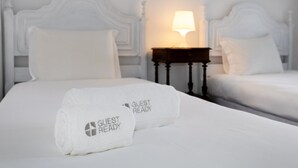 Et hotelværelse med to senge, hvide sengetøj, et natbord med lampe og håndklæder med teksten 'GUEST READY'.