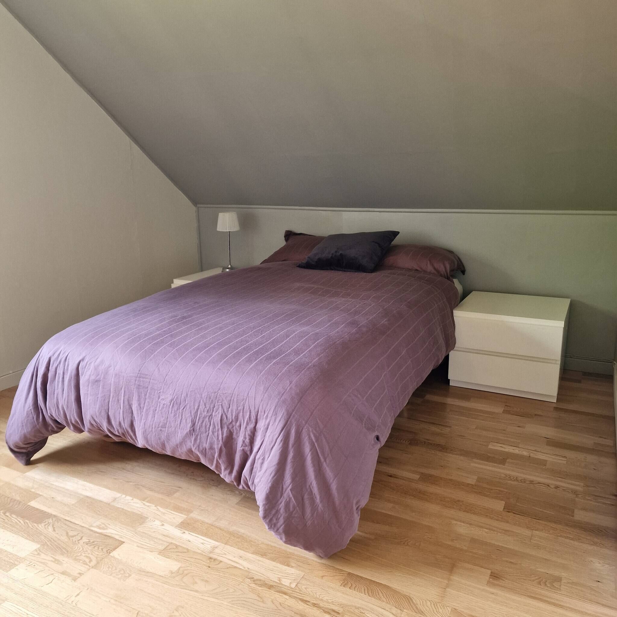 Quarto com cama, mesas de cabeceira, abajur e piso de madeira.