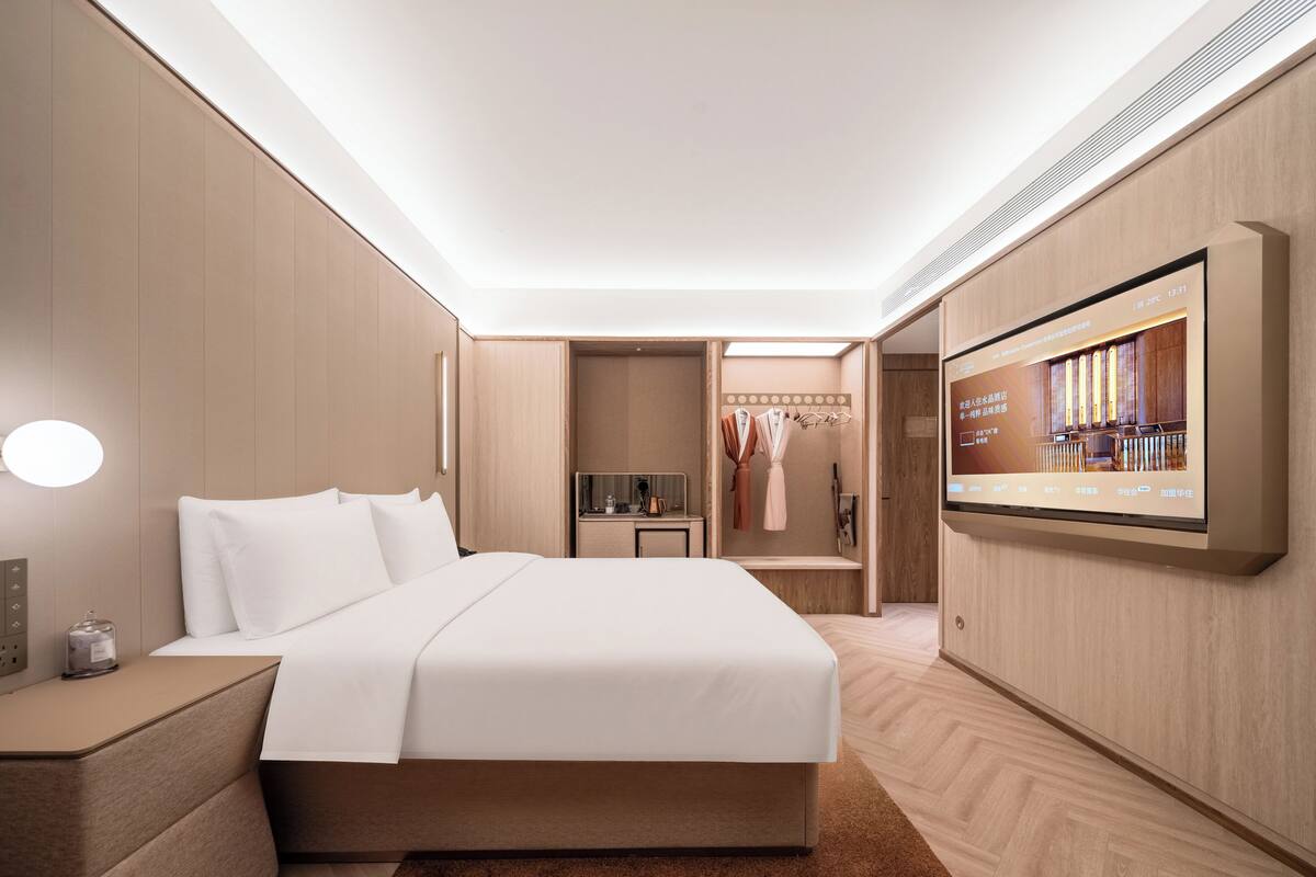 Ein modernes Hotelzimmer mit einem großen Bett, Nachttischen, einem an der Wand befestigten Fernseher und einem begehbaren Kleiderschrank mit Kleidung und Spiegel.