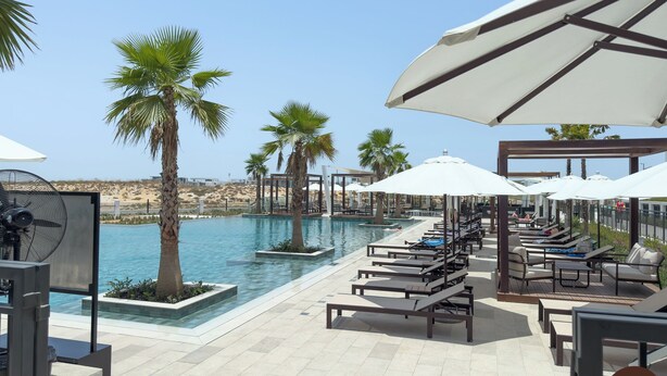 Zona de piscina con sillones de descanso, sombrillas y palmeras.