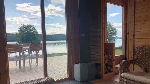 A room with a large glass door leading to an outdoor deck with a table and chairs, a wicker chair, and a fireplace with a log storage.